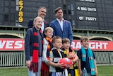 Two men and five children wearing AFL scarfs stand in front of a historic scoreboard