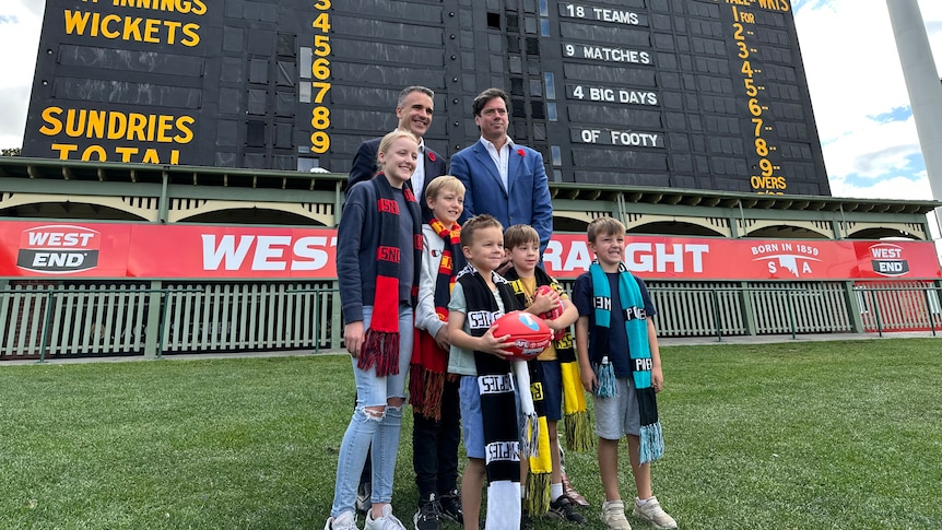 Two men and five children wearing AFL scarfs stand in front of a historic scoreboard