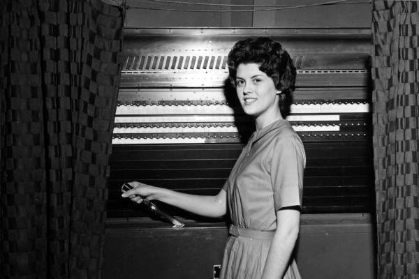 A woman stands with her hand on the lever of a lever voting machine.