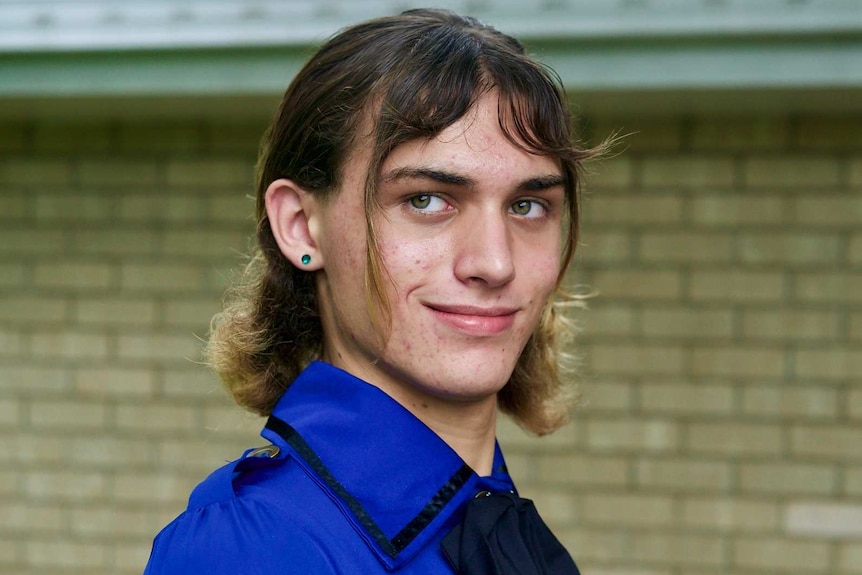 Trans caucasian woman with short brown hair wears blue military-style dress and smiles over her shoulder