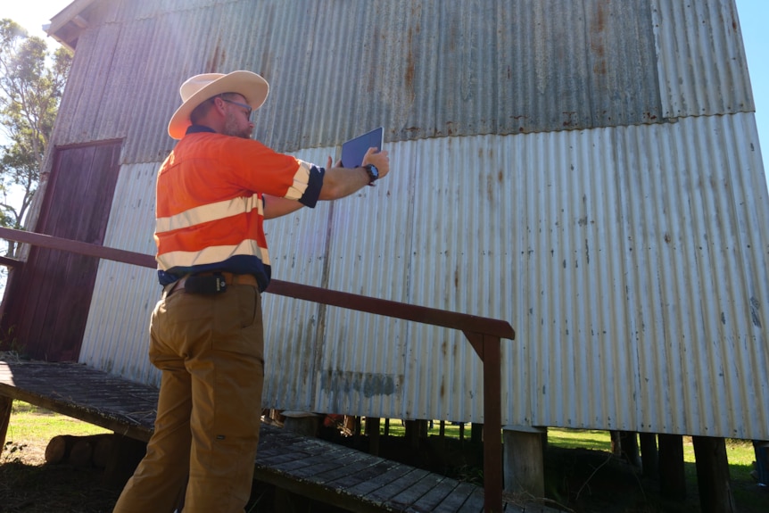 s man in high-vis work clothing holds an ipad up to the exterior wall of an old building