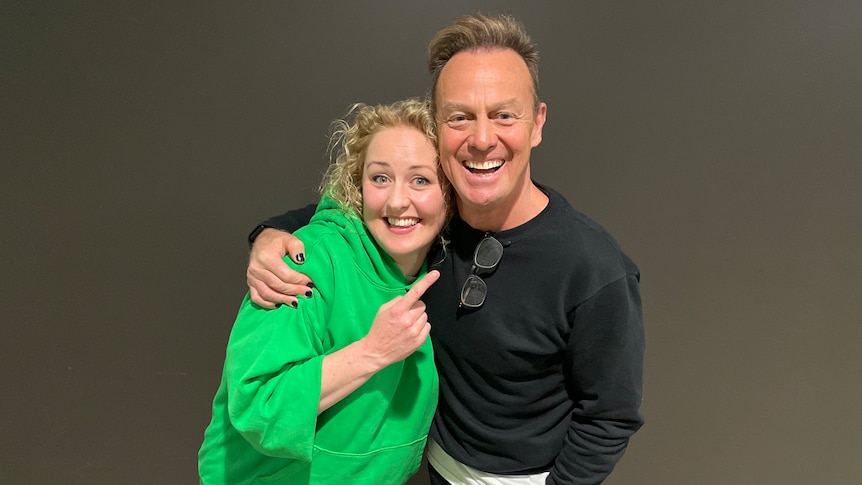 Jason Donovan is in black with his arm around Zan Rowe who is wearing green. They are standing in front of a black wall