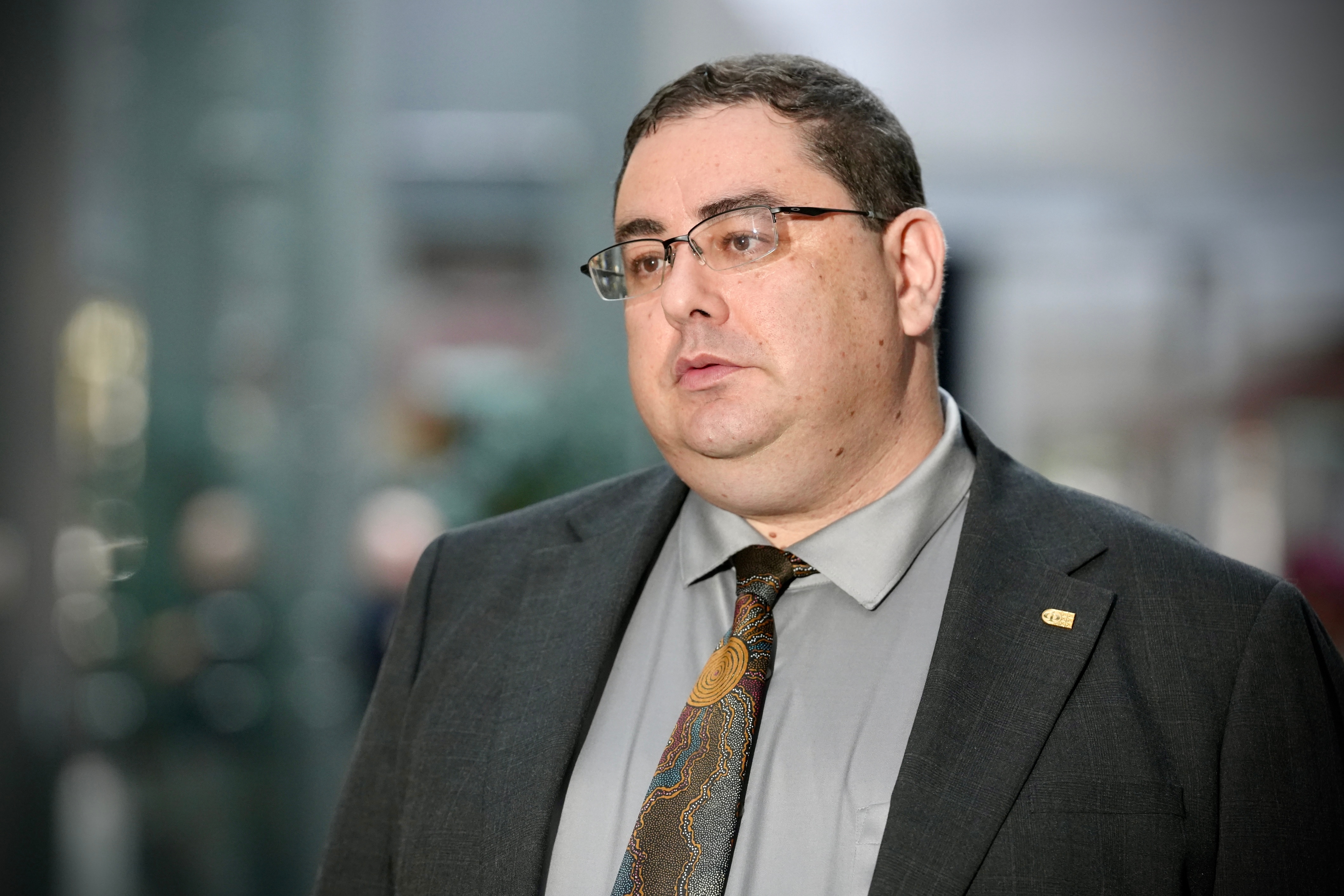 Andre Oboler in a suit, tie, glasses, speaking to a crowd outside an office building.