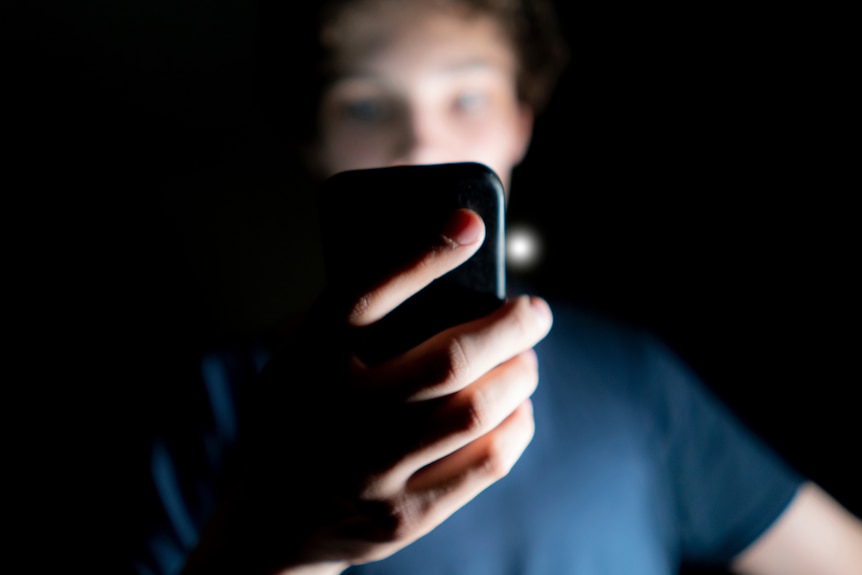 A boy with a blurry face holds a phone, the light illuminating his face against the darkened background.