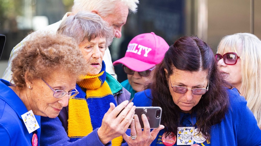 Several mature women gather around one women holding a mobile phone.