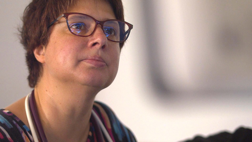 A woman wearing glasses looks up, passed the camera, at a screen.