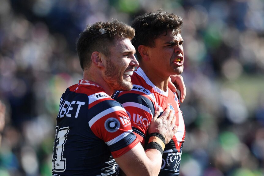 Latrell Mitchell is speaking while a smiling teammate puts his arm over his shoulder and pats him on the chest.