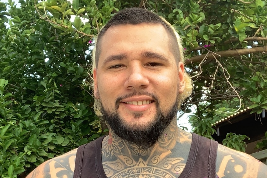 A man with tattoos and facial hair smiles in front of the trees