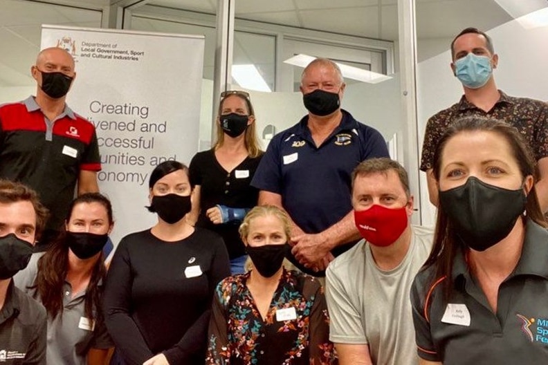 A group of men and women gathered inside, in front of a whiteboard, wearing masks.