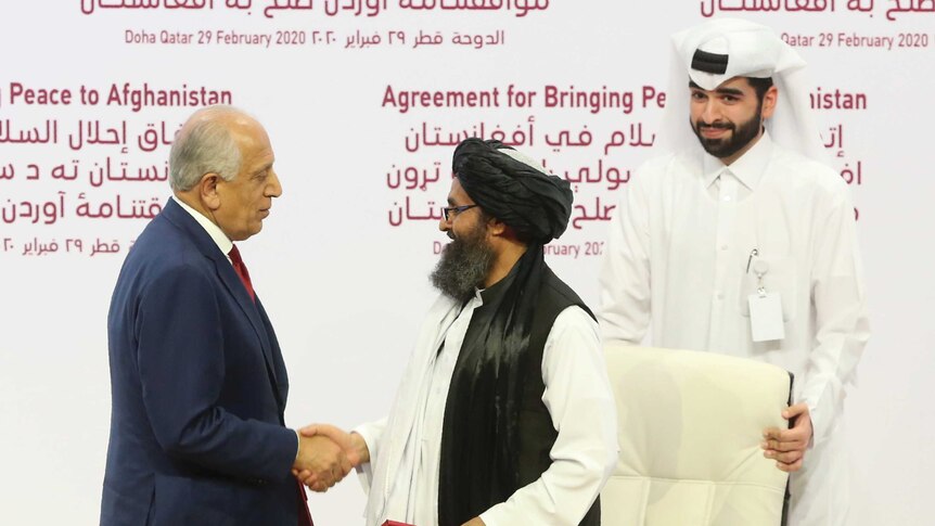 A Taliban wearing a turban and traditional dress shakes hands with a US representatives wearing a suit.