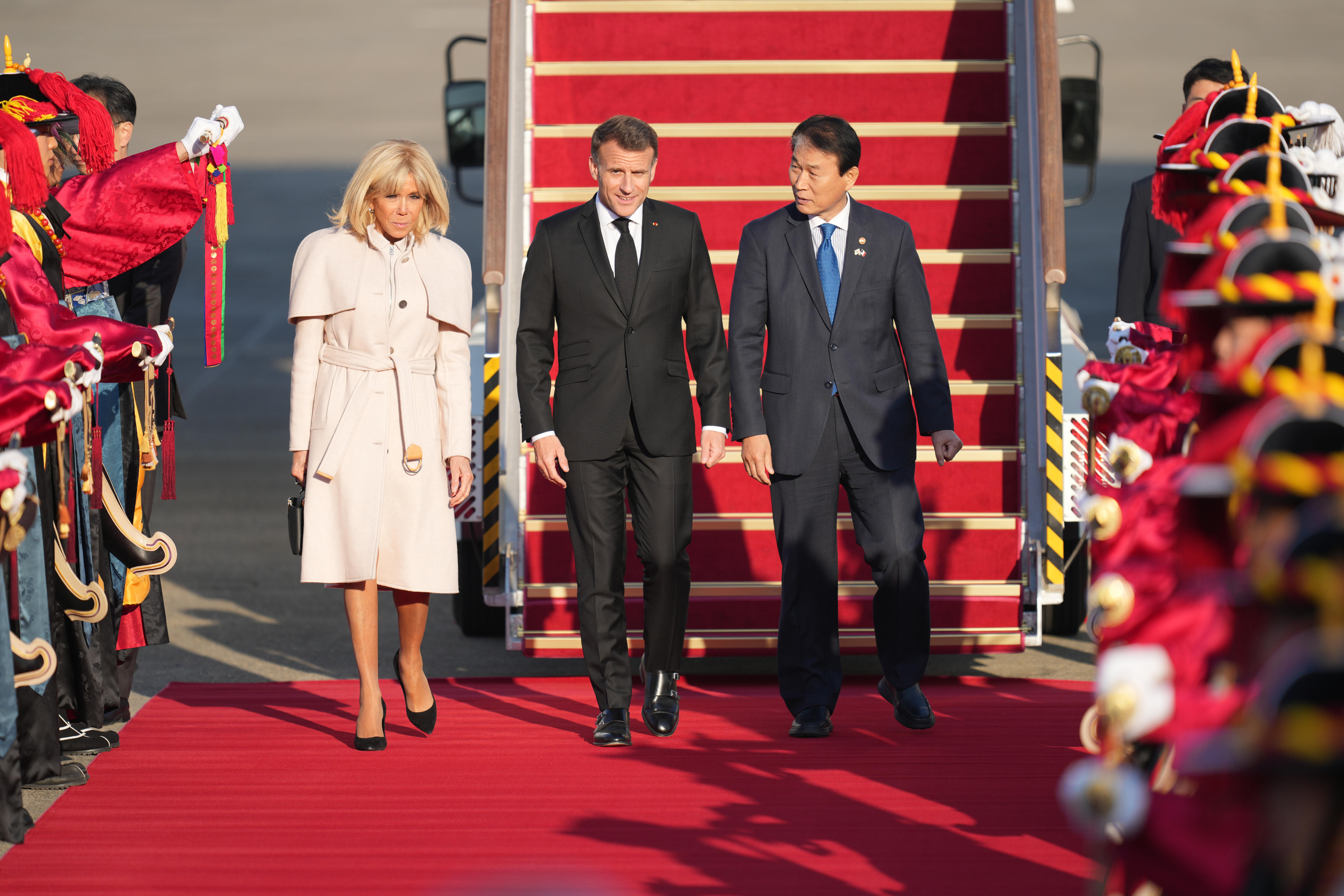 Una mujer y dos hombres caminan uno al lado del otro sobre una alfombra roja a través de una guardia de honor. 