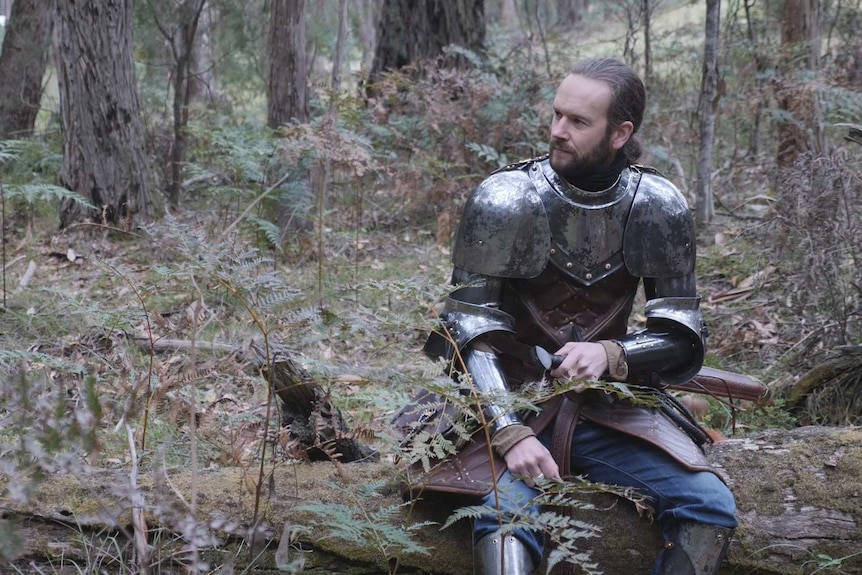A man in a suit of armour sitting on a log in the bush