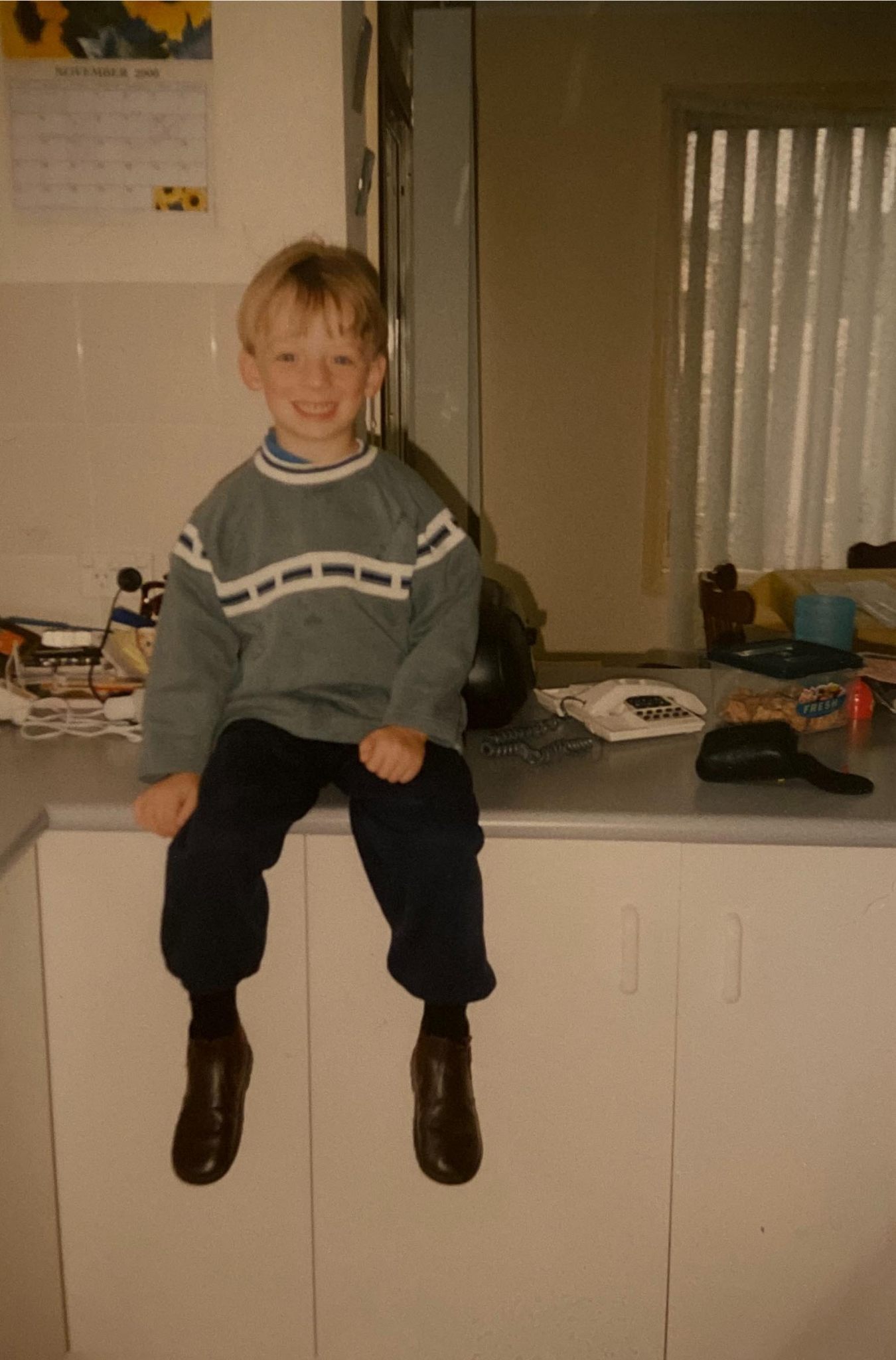Boy with blond hair wearing a jumper sits on a kitchen bench