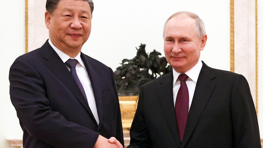 Vladimir Putin, dressed in a suit, shakes hands with Xi Jinping, in a suit, in a room in Moscow.