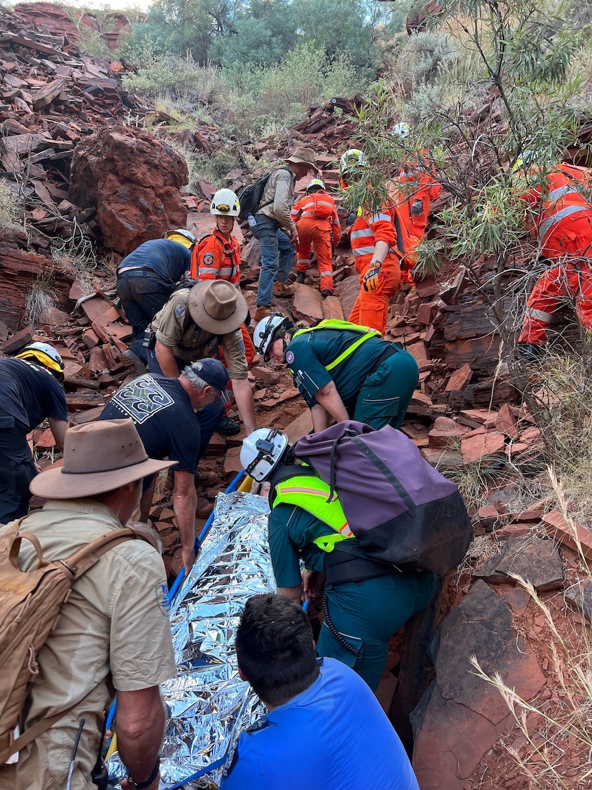 A team of people carrying a woman on a stretcher up a rocky, steep hill.