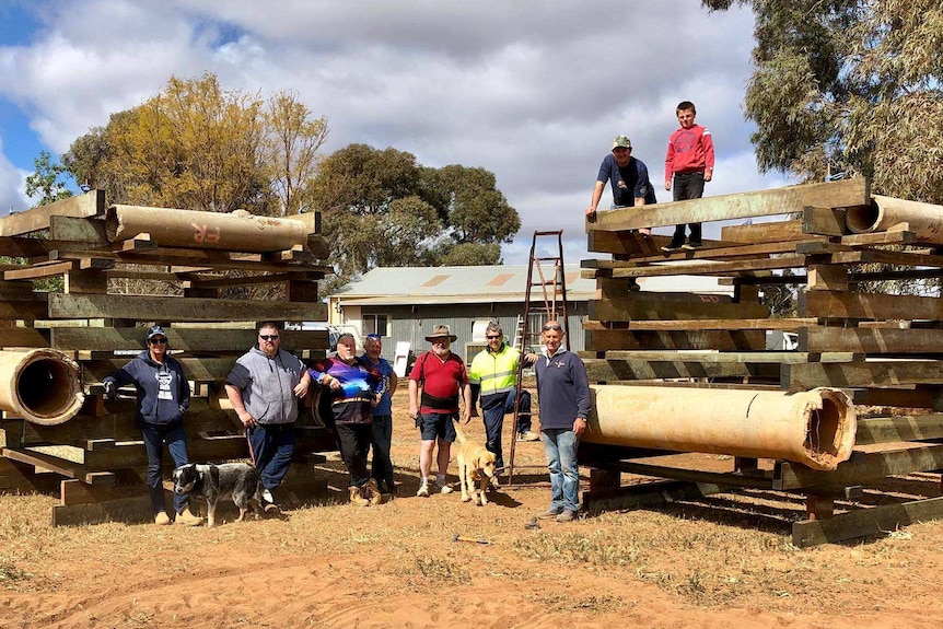 A group of people pose next to two giant cube-shaped wooden structures built from various sized wood slabs and tubes