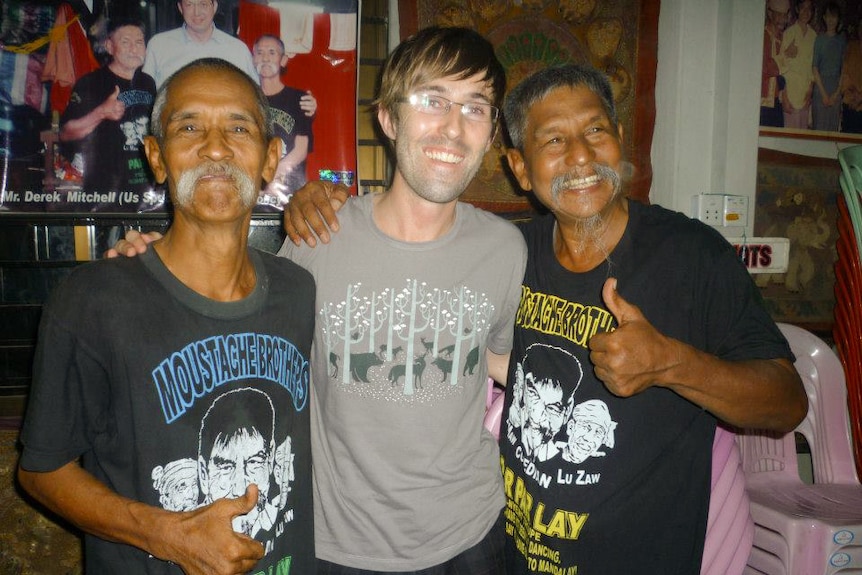 A man wearing glasses and a grey shirt stands beside two men with moustaches and wearing black shirts.