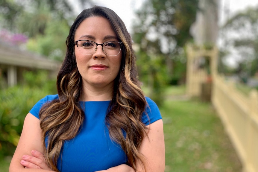 A woman with glasses and a blue top looking at camera, arms folded