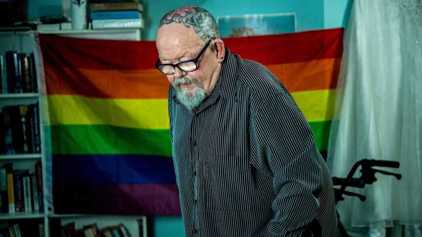 Geoffrey Ostling stands up in front of a rainbow flag in his aged care room.