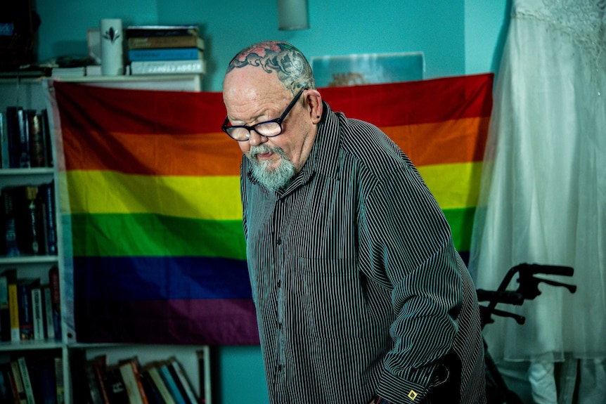 Geoffrey Ostling stands up in front of a rainbow flag in his aged care room.