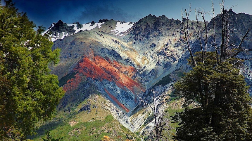 View of craggy mountains rising from lush green slopes, with rocky hues, snow-capped peaks and areas of striking orange.