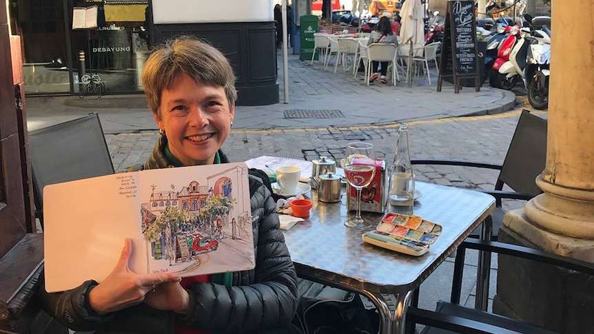 An artist displays a sketch of on a Spanish street in front of her where she is sitting at a table with a drink.