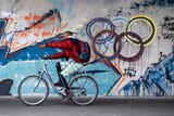 A man on a bike rides past a wall, with a painting of the olympic rings on it.