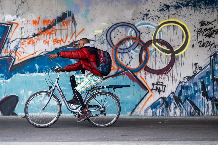 A man on a bike rides past a wall, with a painting of the olympic rings on it.