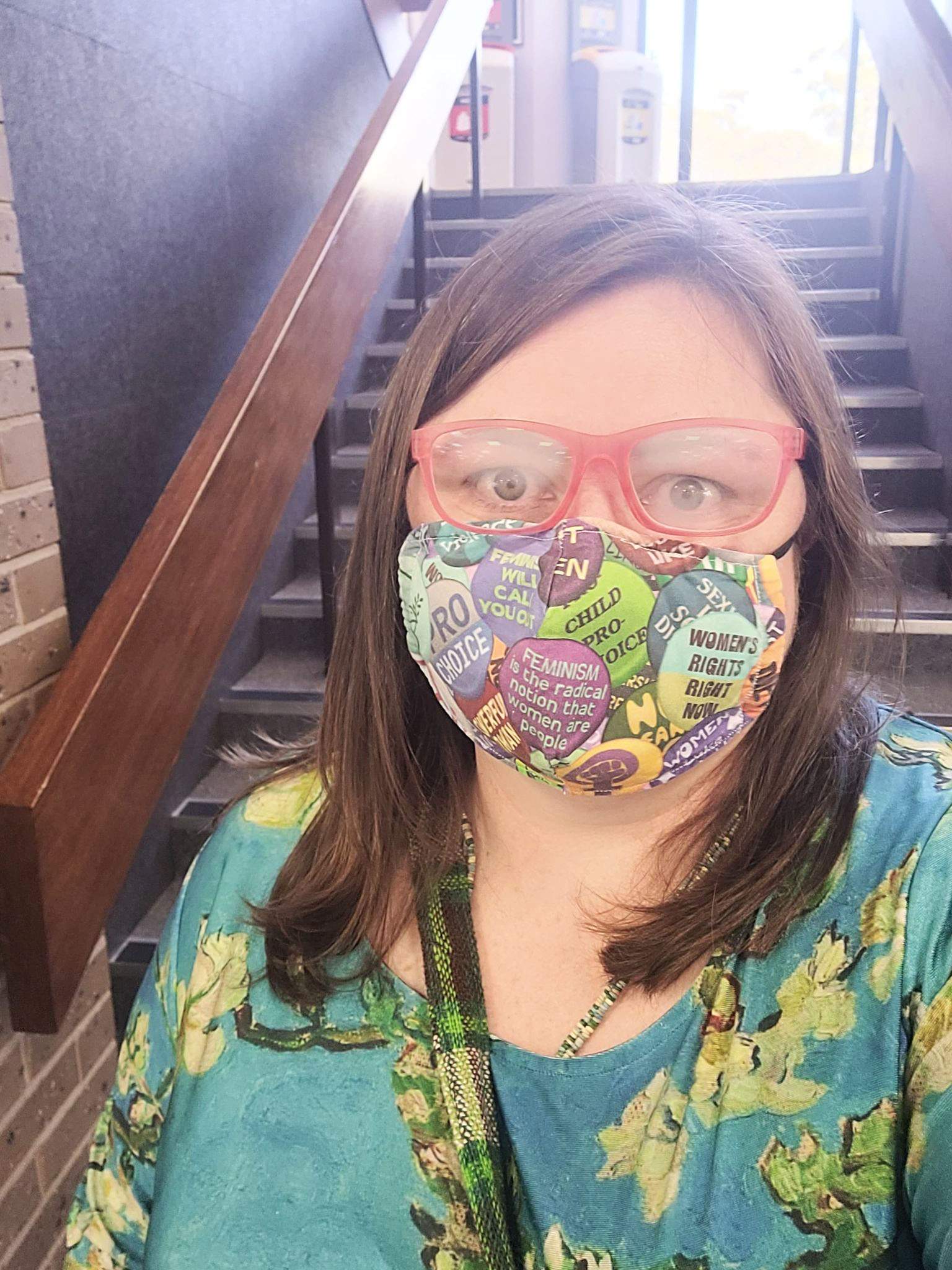 Mid level shot of a woman wearing a mask standing in front of flight of stairs, she is wearing a mask and glasses.