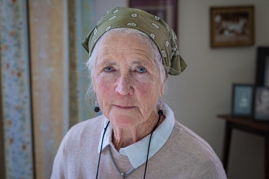 An old woman wearing a beige jumper and green bandana stares into the camera.