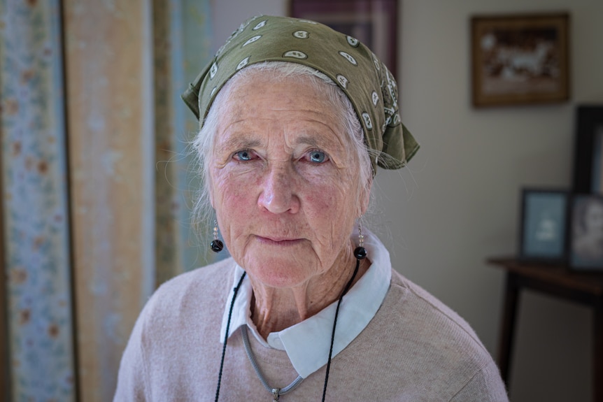 An old woman wearing a beige jumper and green bandana stares into the camera.