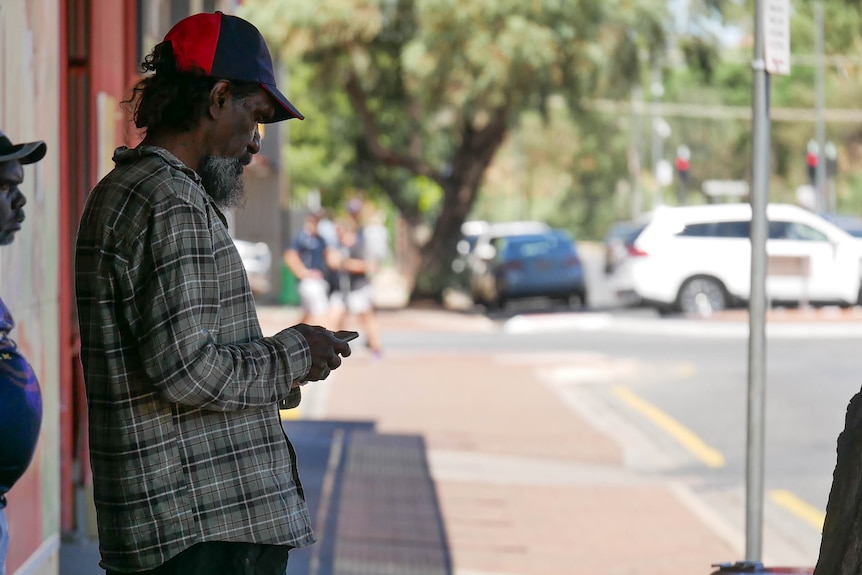 An Aboriginal man leans against a wall while looking at his phone in Alice Springs.