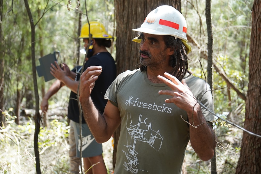 A man in a hard hat explains something in the bush using his hands expressively.