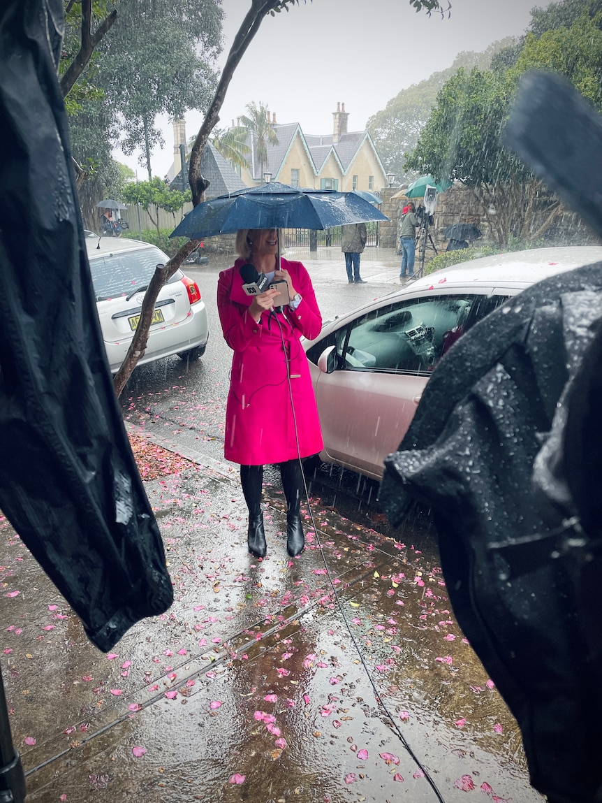 Woman standing under umbrella in front of TV camera as rain pours down.