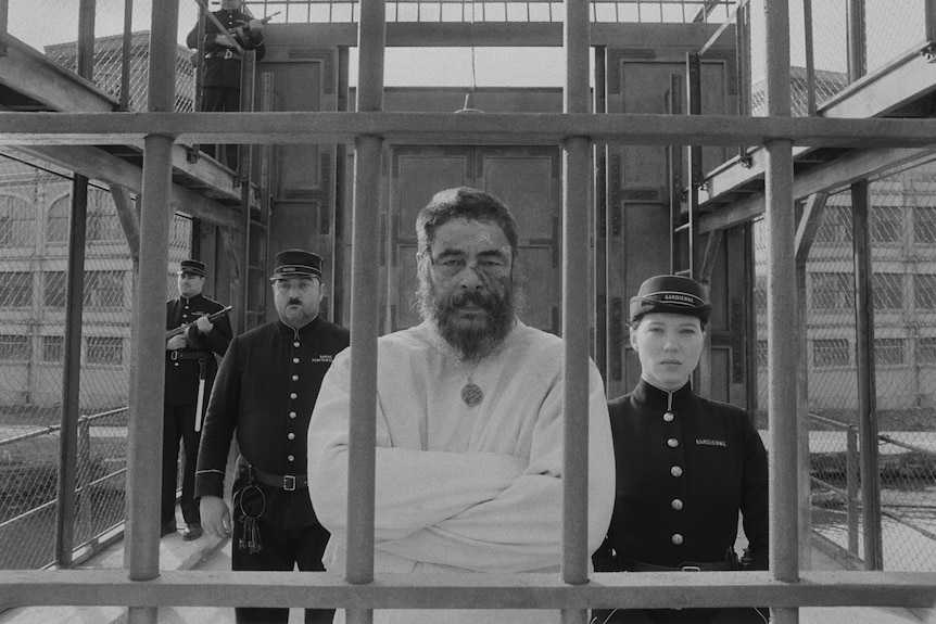 In B&W, a bearded 50-something Hispanic man in a straitjacket is behind bars, flanked by prison officers, including a woman
