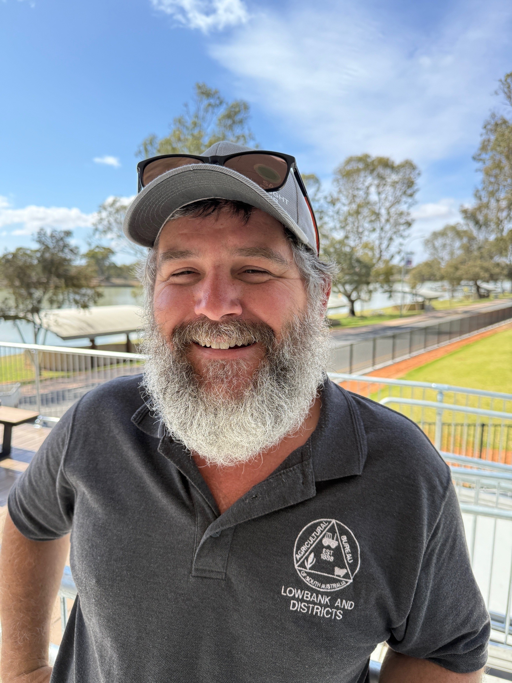 A smiling man with a bushy grey beard stands with the Murray River behidn him.