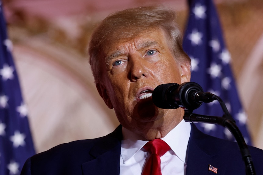 Donald Trump speaks closely into a microphone with two American flags behind him.