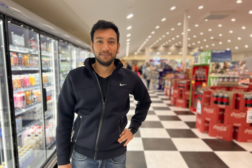 a dark haired olive skinned man stands in the middle of a supermarket aisle