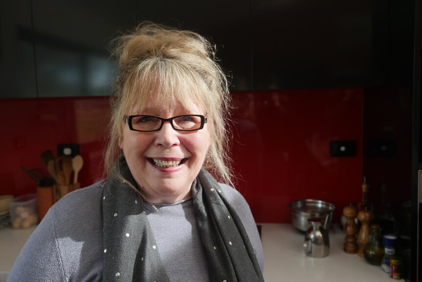 A bespectacled woman with blonde hair in a bun smiling in a kitchen.