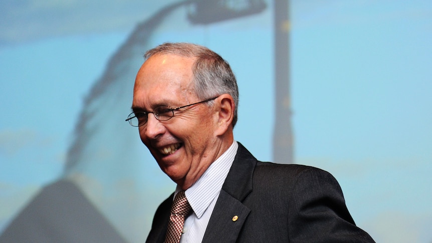 A man, wearing a suit and glasses, laughing in front of a picture of a coal pile