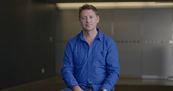 A man in his mid-forties sits, wearing a blue shirt, infront of a glass backdrop.