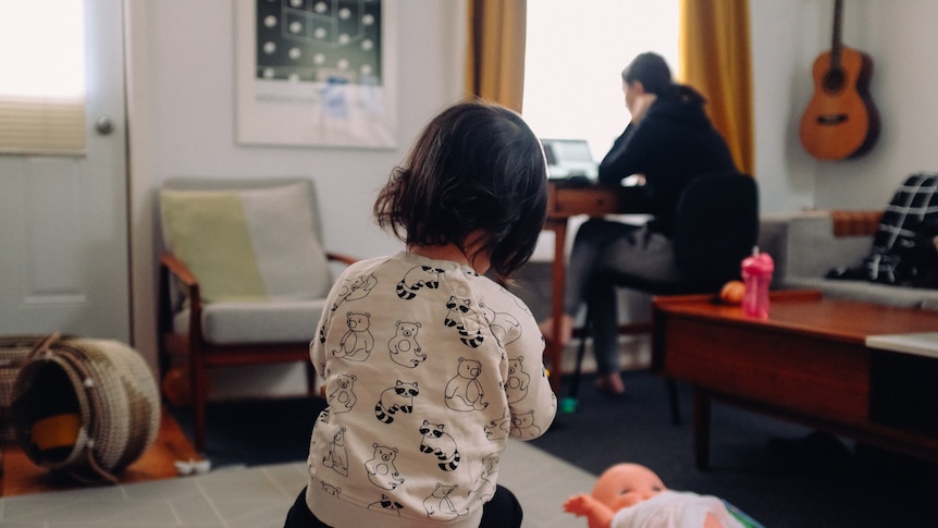 Image focuses on a young child playing by herself. An adult sits at a computer in the background.