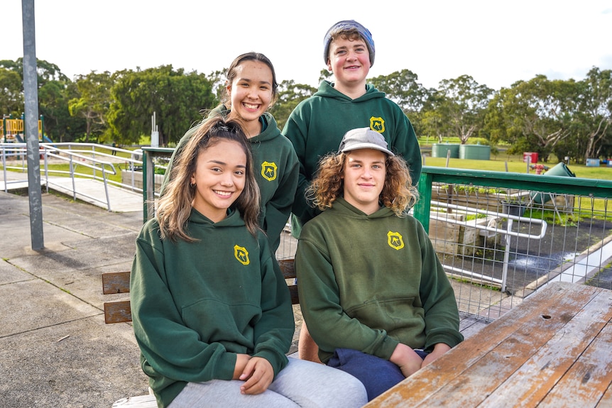 Four teenage students in green school uniforms sit outside at a school bench together smiling.
