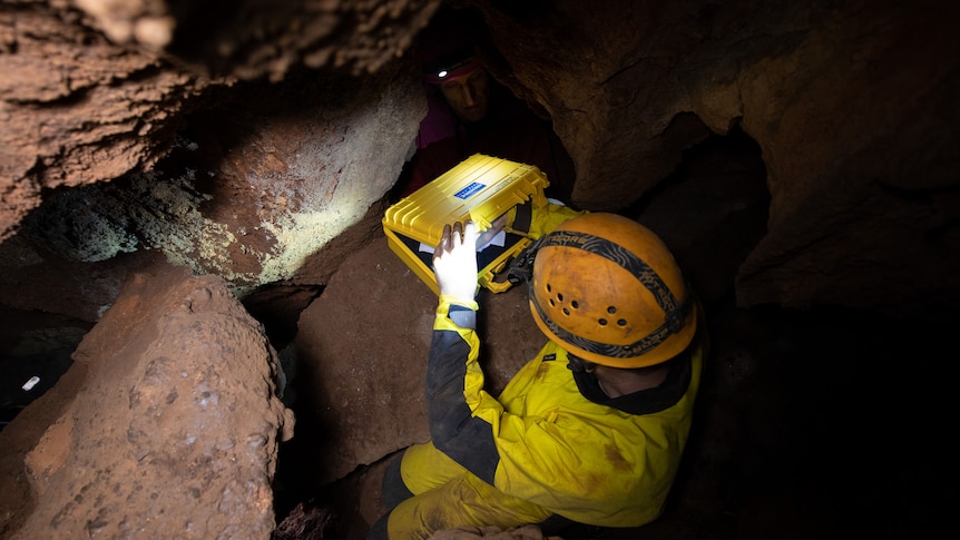 A man in caving gear puts something into a yellow briefcase. Out of the dark there's a second face looking up 