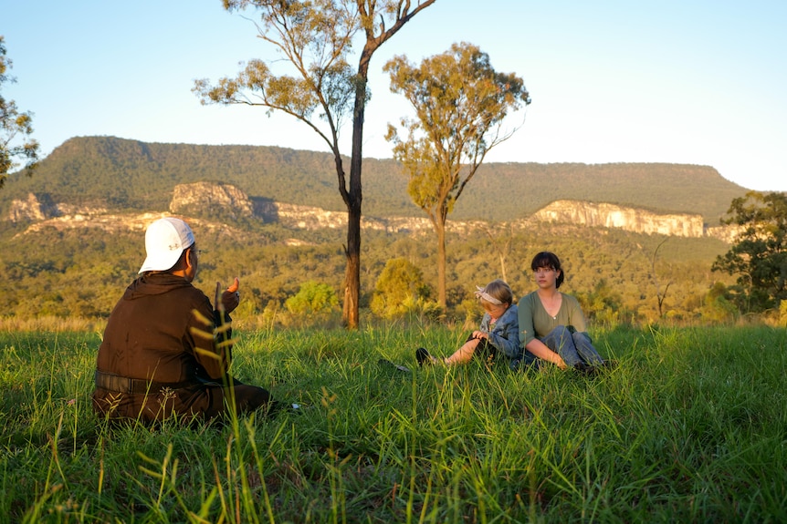 Melissa and two girls sitting in long grass, sandstone cliffs and trees behind.