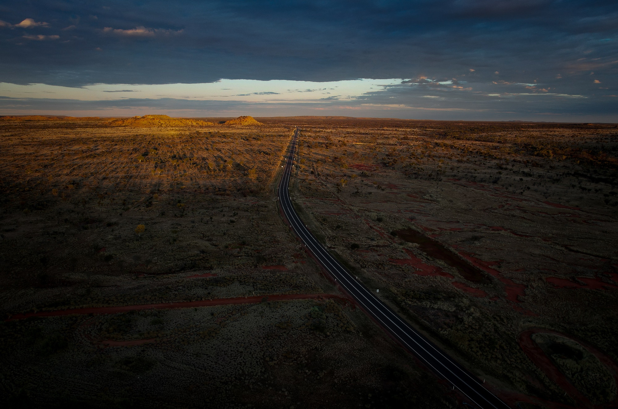 Aerial shot over vast outback landscape shows a dark bitumen highway running through the orange dirt, dotted with green trees.