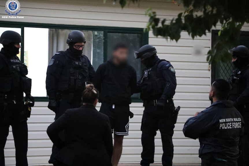 A group of heavily armed police officers surround a man out the front of a house.