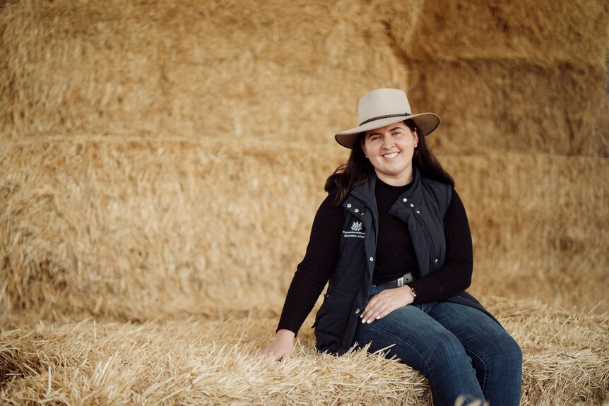 Image of a young woman wearing a black bodywarmer and a hat while sitting on bales of hay.