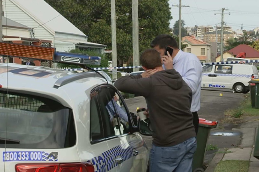 Two detectives lean on a police car while one of them talks on the phone