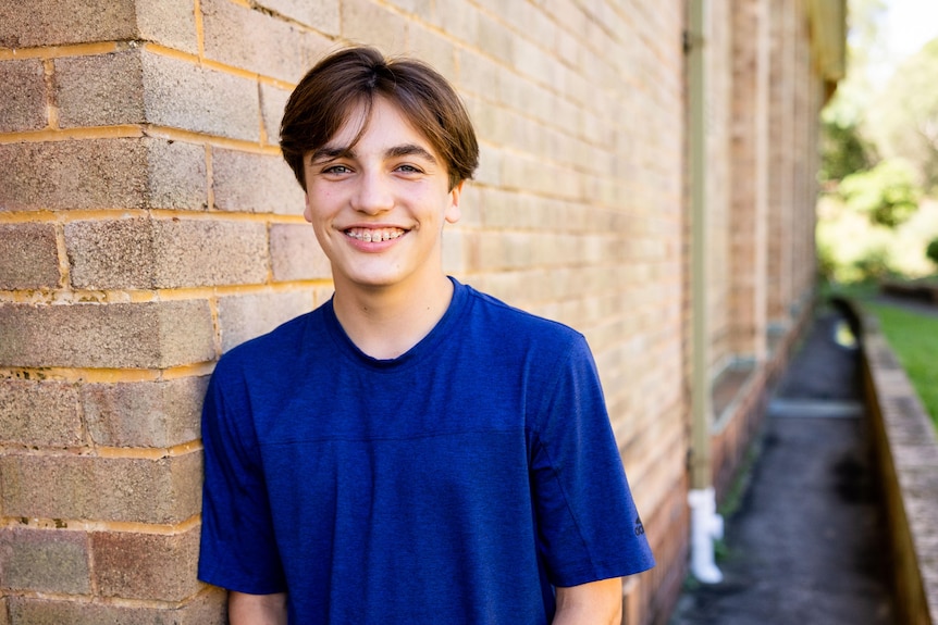 A teenage boy in a blue shirt with braces smiles widely while leaning against a brick wall.